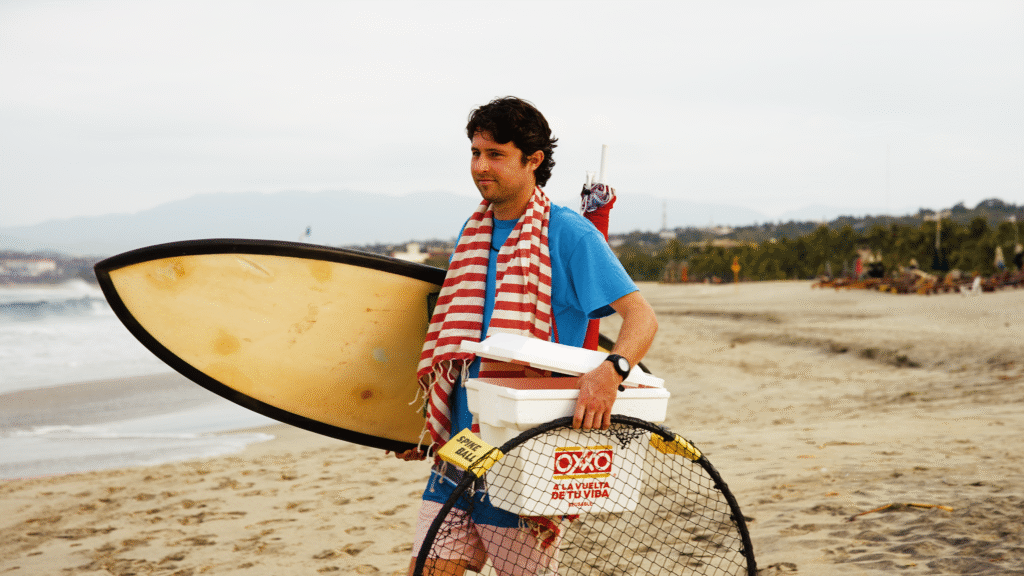 Mack Linstead, Beach Sherpa Founder, carrying a surfboard, cooler, spikeball set up, towel, and umbrella on the beach.
